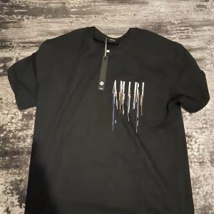 Svart t-shirt från Amiri med färgstänkt logga - Säljer en svart t-shirt från Amiri med tryckt logga och färgstänk i flera färger både fram och bak. T-shirten har rund hals och normal passform. Perfekt för dig som gillar streetwear och vill sticka ut.