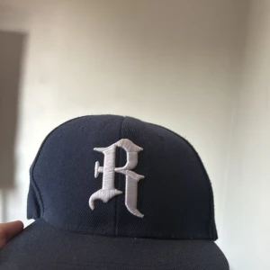Baseball keps - Snygg mörkblå keps med ett stort vitt broderat R framtill och färgglad brodyr på sidan. Justerbar snapback baktill för perfekt passform. Klassisk streetstil som passar till det mesta.
