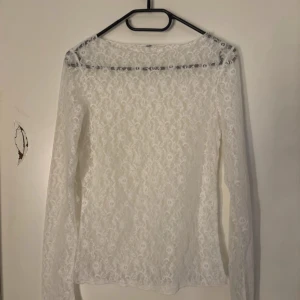 Vit spets topp  med lång ärm - Superfin vit topp i spets med långärmad design och transparent känsla. Blusen har ett romantiskt blommönster i spetsen och en rak passform. Perfekt för dig som vill ha en söt och stilren look.