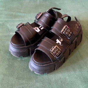 Buffalo chunky svarta sandaler med spännen - Buffalos, svarta chunky sandaler med breda remmar och metallspännen. Grov plattformsula och öppen tå ger en edgy vibe. Remmarna har coola nitar och öglor för extra detalj. Perfekta för dig som gillar statement-skor och vill sticka ut i sommar.
