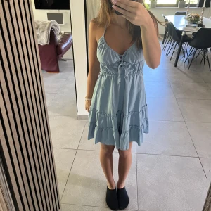 Ljusblå kort klänning med volanger - Supersöt ljusblå kort klänning med volanger och knytning framtill. Klänningen har smala axelband som är knutna runt nacken med öppen rygg. 