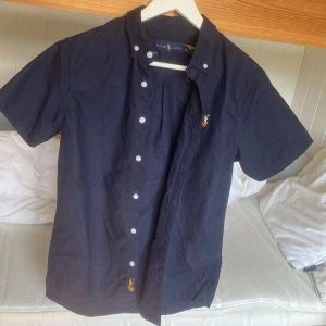 Mörkblå kortärmad skjorta Ralph Lauren - Snygg mörkblå skjorta från Ralph Lauren med korta ärmar och klassisk button-down krage. Skjortan har broderad logga på bröstet och vita knappar framtill. Tillverkad i mjuk bomull, perfekt för en clean och stilren look.