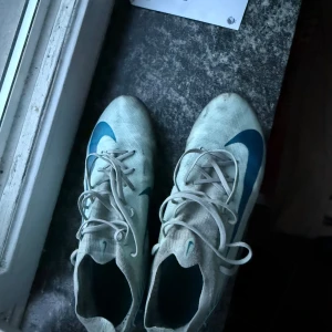 Nike vita fotbollsskor med blå swoosh - Säljer ett par vita Nike fotbollsskor med blå swoosh på sidorna. Skorna har snörning och dobbar under sulan för bra grepp på planen!PRISET KAN DISKUTERAS!!!