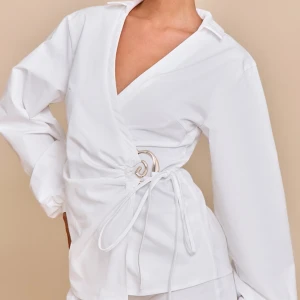 Vit omlottblus med knytband - Snygg vit omlottblus med v-ringning och långa ärmar. Blusen har knytband i midjan och en dekorativ metallring på sidan