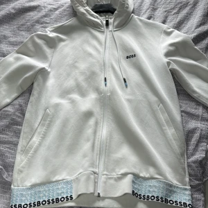 Vit hoodie från BOSS med dragkedja - Snygg vit hoodie från BOSS med hel dragkedja, huva med snörning och diskret BOSS-logga på bröstet. Nedtill finns en cool detalj med upprepad BOSS-text i svart och blått. Perfekt för en clean och stilren look.