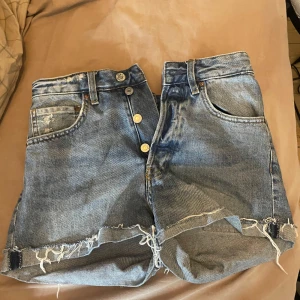 Blå jeansshorts med fransig kant - Snygga blå jeansshorts med hög midja och fransig kant nertill. Slitna detaljer och knappar framtill. Perfekta för sommaren!☀️