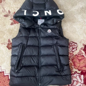 Svart dunväst från Moncler med huva - Snygg svart dunväst från Moncler med stor huva och vit logotyptext på kanten. Västen har två dragkedjefickor och klassisk Moncler-logga på bröstet. Perfekt för lager-på-lager och streetstyle. Storlek S