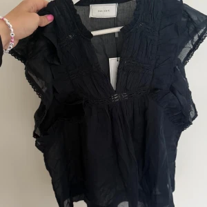 Mörkblå blus med volang från Neo Noir - Supersöt mörkblå blus från Neo Noir i luftig bomull. Modellen har v-ringning, volangdetaljer och lätt transparent tyg. Perfekt för dig som gillar en romantisk och trendig look med extra detaljer vid axlar och hals.