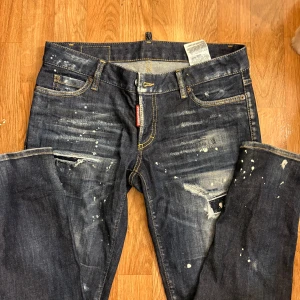 Mörkblå Dsquared2 jeans med slitningar - Snygga mörkblå jeans från Dsquared2 med coola slitningar och färgstänk för en edgy look. Klassisk femficksmodell med kontrastsömmar och Dsquared2-logga vid fickan. Jeansen har en rak passform och är tillverkade i robust denim.