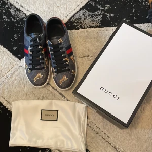 Gucci sneakers med tigerprint - Snygga Gucci sneakers med blå bas, röda och marinblå ränder på sidan och coolt tigerprint över hela skon. Skorna har svarta skosnören och vit sula. Perfekta för dig som vill sticka ut med lyxig streetstyle.