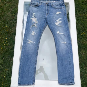 Jeans - Säljer ett par &Denim jeans med kraftigt distressed design (designade hål och slitage). Modell: Tapered Low Waist Dropped Crotch. Storlek W33 L34. Tapered fit med låg midja. Jeansen är i bra skick utifrån att slitaget är en del av designen. Bara att höra av dig vid minsta fundering!