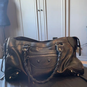 Svart skinnväska med flätade handtag - Snygg svart handväska i skinn med flätade handtag och silverfärgade metalldetaljer. Väskan har en dragkedja upptill, framficka med dragkedja och snygga nitar. Perfekt storlek för att få plats med allt du behöver och passar till många olika stilar.