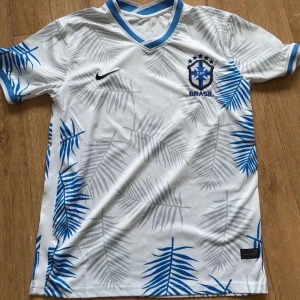Vit Brasilien fotbollströja Nike S - Vit Brasilien fotbollströja från Nike med blå och grå palmbladsmönster. Tröjan har blå detaljer vid krage och ärmslut, CBF-emblem på bröstet och Nike-logga. Tillverkad i lätt och ventilerande Dri-FIT-material, perfekt för träning eller match.