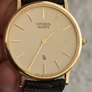 Citizen Quartz guld klocka med läderband - Stilren och klassisk Citizen Quartz klocka med rund guldfärgad boett och ljus urtavla. Tunt, elegant armband i mörkbrunt läder med struktur. Minimalistisk design med guldiga index och visare. Perfekt för dig som gillar tidlös och clean stil. Behöver nytt batteri, fullt fungerande, repfritt glas, diameter på klockan är ca 31 mm  märken finns på armbandet, 