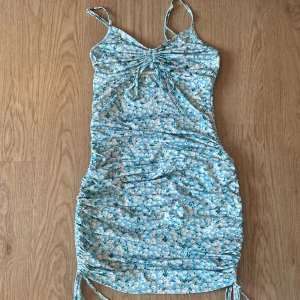 Blommig blå kortklänning från SHEIN storlek XS - Supersöt blå kortklänning från SHEIN med vita och gula blommor. Klänningen har smala axelband, snörning och cut-out vid bysten samt rynkade sidor med knytband nertill. Perfekt för sommarens alla tillfällen!