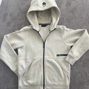 Beige huvtröja från Sail Racing - Säljer en snygg beige huvtröja från Sail Racing. Den har en dragkedja framtill och en mysig huva med ett coolt märke på. Perfekt för höst och vår, med långa ärmar och en skön passform. Tröjan är i bra skick och redo för nya äventyr!