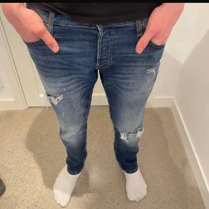 Blå slitna jeans med raka ben - Snygga blå jeans med slitningar och hål på knäna. Jeansen har en klassisk femficksmodell, normal passform och raka ben. Materialet är jeans med en tvättad look och ljusare partier framtill. Perfekta för en avslappnad och trendig stil.