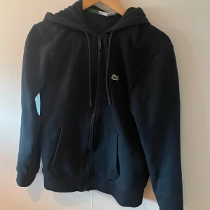 Svart hoodie från Lacoste med dragkedja - Säljer denna svarta Lacoste zip hoodie. Nypris ca 1300kr. Använd 3 gånger, säljer då den är för liten för mig. Inga tecken på användning. 
