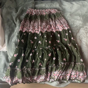 Grön blommig långkjol med volanger - Supersöt långkjol i mörkgrönt med rosa och vita blommor, köpt utomlands. Kjolen har ett insytt resårband i midjan, ser väldigt liten ut men kan stretchas ut mycke kjolen har även flera volanglager som ger en flowy känsla. Perfekt för dig som gillar boho-stil och vill ha något bekvämt och snyggt till sommaren.