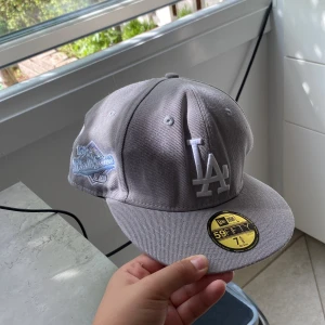 Grå LA Dodgers keps New Era 59FIFTY - Snygg grå LA Dodgers keps från New Era 59FIFTY-serien. Klassisk modell med broderad LA-logga framtill och patch på sidan. Rak skärm och strukturerad passform. Perfekt för dig som gillar streetwear och sportig stil.