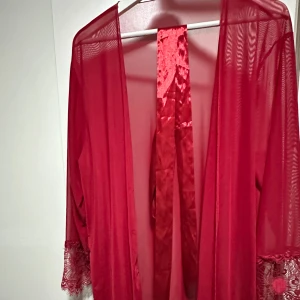Röd transparent omlottkofta med spets - Snygg röd transparent omlottkofta med breda spetsdetaljer vid ärmslut och nederkant. Koftan har ett matchande rött band i satin att knyta med i midjan. Perfekt för dig som vill ha något extra och unikt i garderoben.