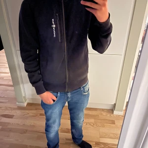 Svart hoodie från Sail Racing med dragkedja - Svart hoodie från Sail Racing med hel dragkedja framtill och huva. Tröjan har en bröstficka med dragkedja och logga på vänster sida. Den är solblekt.