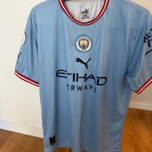 Manchester City De Bruyne matchtröja - Manchester City matchtröja med De Bruyne 17 på ryggen. Ljusblå med röda och vita detaljer vid krage och ärmslut. Puma-logga, klubbmärke och Etihad Airways-tryck på bröstet. Korta ärmar, tillverkad i lätt och ventilerande material.