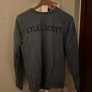 Blå sweatshirt från Lyle & Scott - Snygg blå sweatshirt från Lyle & Scott med stort tryck på ryggen. Tröjan har rund halsringning och långa ärmar, perfekt för en chill och stilren look. 
