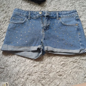 Jeansshorts - Snygga blå jeansshorts med uppvikta ben och coola silvriga nitar framtill. Klassisk femficksmodell med bälteshällor och bakfickor. Perfekta för sommardagar när du vill ha en chill och trendig look.