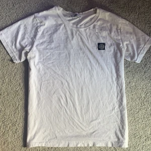 Vit Stone Island t-shirt med patch - Vit t-shirt från Stone Island med klassisk rund hals och korta ärmar. På bröstet sitter en ikonisk Stone Island-patch i svart och vitt. T-shirten är i mjuk bomull och har en clean, stilren look som passar perfekt till jeans eller shorts.