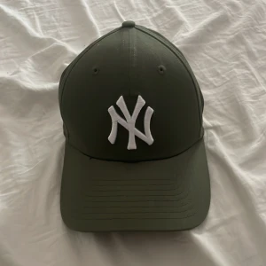 Olivgrön New Era Yankees keps - Snygg olivgrön New Era 39THIRTY keps med klassiskt New York Yankees broderat vitt logotyp framtill. Kepsen har böjd skärm och stretchig passform. Tillverkad i bomull och polyester, perfekt för en sportig och stilren look.
