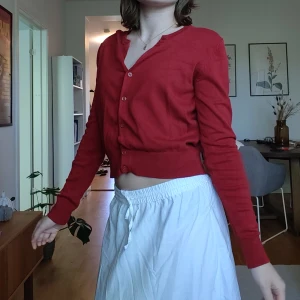 Röd kofta från H&M - Röd kofta från H&M med långa ärmar och knappar framtill. Klassisk modell med ribbade muddar vid ärmslut och nederkant. Perfekt att slänga över en topp eller klänning för en extra färgklick.