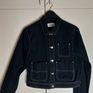 Svart jeansjacka med vita detaljer från Weekday - Svart jeansjacka från Weekday med kontrasterande vita sömmar, klassisk krage och tre stora fickor framtill. Jackan har raka linjer, knäppning med silverfärgade knappar och är långärmad. Perfekt för dig som gillar minimalistisk och stilren look.