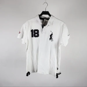 Vit polo t-shirt från CNK Bike Polo - Vit polo t-shirt från CNK Bike Polo med svart tryck fram och bak, nummer 18 och 91 samt texten 'Bike Polo'. Klassisk krage och korta ärmar. Tillverkad i 100% bomull, perfekt för en sportig och avslappnad stil.