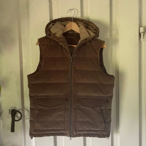 Brun manchesterdunväst från Stenströms - Snygg brun dunväst från Stenströms i manchester med quiltad look och avtagbar huva. Västen har två stora fickor framtill och dragkedja hela vägen. Perfekt för lager-på-lager till sommarkvällar. Otroligt eftertraktad modell. Använd i 3 månader utan slitage ser som ny ut. Vid frågor och funderingar är det bara att höra av sig!😊                                                                                                                            Mvh Doré.reselll