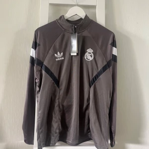 Adidas Real Madrid långärmad tröja L - Snygg långärmad Real Madrid tröja från Adidas i mörkgrått med svarta och vita detaljer. Tröjan har half zip, hög krage och klubbmärke på bröstet. Perfekt för fotboll eller chill. Tillverkad i polyester för en sportig känsla.