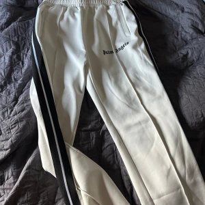 Palm Angels byxor  - Snygga vita mjukisbyxor från Palm Angels med svarta ränder längs sidorna och broderad logga på låret. Byxorna har elastisk midja och är gjorda i bomull och polyester för en skön och sportig känsla. Perfekta för en chill och trendig look.  INTE ANVÄNDA!