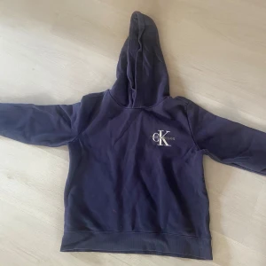 Mörkblå Calvin Klein hoodie - Mörkblå hoodie från Calvin Klein Jeans med klassisk logga broderad på bröstet. Tröjan har huva och långa ärmar, tillverkad i mjuk bomull. Perfekt för en chill och avslappnad stil.den har en liten fläck på magen.