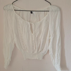 Vit blus med spets från H&M Divided - Superfin vit blus från H&M Divided med långa ärmar, smockad midja och spetsdetaljer framtill. Blusen har knytning vid halsen och är gjord i ett lätt och luftigt material. Perfekt för en romantisk och bohemisk look.