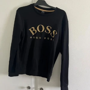 Hugo Boss sweatshirt - Svart långärmad tröja från Hugo Boss med guldig logga framtill. Klassisk rund halsringning och normal passform. Perfekt för dig som gillar stilrena och snygga plagg med tydlig branding. Pris går att diskuteras via snabb affär. Storlek L men kan passa M