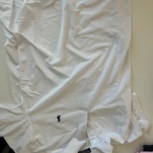 Vit skjorta från Polo Ralph Lauren - Klassisk vit skjorta från Polo Ralph Lauren med broderad logga på bröstet. Skjortan har knappar framtill och är tillverkad i bomull. Perfekt för en clean och stilren look.