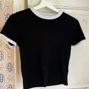 Svart ribbad topp med vita detaljer - En svart ribbad topp med korta ärmar och kontrasterande vita muddar vid halsringning och ärmslut. Enkel och stilren design som passar perfekt till jeans eller kjol. T-shirten har en tight passform och är superenkel att matcha.