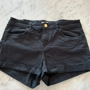 Svarta shorts med fickor och knapp - Svarta shorts med klassisk femficksdesign, bälteshällor och guldfärgad knapp framtill. De har uppvikta benslut och är sydda i ett mjukt bomullstyg. Perfekta för varma dagar och enkel att matcha med allt.