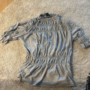Grå smockad blus med puffärm - Supersöt grå blus med smockade detaljer över bröst, midja och ärmslut. Blusen har korta puffärmar och en hög halsringning. Perfekt för dig som gillar en feminin och trendig look. Mjuk och skön i bomullsmix.