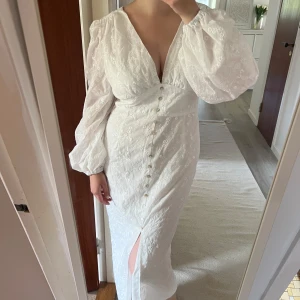 Vit långklänning med broderade blommor - Superfin vit långklänning med broderade blommor, v-ringning och puffiga långärmar. Klänningen har knappar framtill och en slits som ger extra edge. Perfekt för sommarens alla tillfällen när du vill känna dig extra dreamy. Dragkedja där bak 