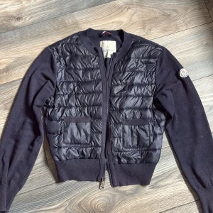 Svart Moncler cardigan jacka med dun - Snygg svart cardigan jacka från Moncler med quiltad dunfront och stickade ärmar. Jackan har två framfickor, ribbade muddar och dragkedja framtill. Perfekt för dig som vill ha en stilren och sportig look med exklusiv känsla.
