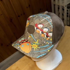 Gucci x Disney keps med Kalle Anka - Snygg keps från Gucci x Disney med brunt GG-mönster och färgglada tryck av Kalle Anka och Knatte, Fnatte och Tjatte i rymden. Kepsen har mesh på sidorna och justerbar rem bak. Perfekt för dig som vill sticka ut med en unik accessoar.