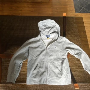 Grå hoodie från Polo Ralph Lauren - Snygg ljusgrå hoodie från Polo Ralph Lauren, Storlek L i barnstorlek (14-16). Säljer denna snygga hoodien på grund av att jag vuxit ur den, skick 7-8/10 använd många gånger, ganska urtvättad och några få fläckar men de är knappt synliga. Priset kan diskuteras.