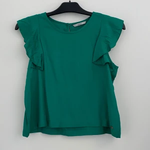 Grön blus med volang från Tiffosi - Snygg grön blus från Tiffosi med volangdetaljer på axlarna. Ärmlös modell med rund halsringning och knapp i nacken. Använd fåtal gånger. Storlek: 164.   Hör av dig vid frågor!                                           Köparen står för frakten. 