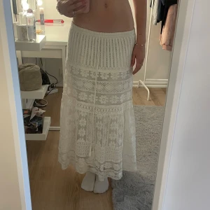 Vit långkjol med spets och snörning - Superfin vit långkjol med broderade spetsdetaljer och blommigt mönster. Kjolen har snörning i midjan och är tillverkad i ett luftigt material som känns perfekt för sommaren. Varan har en fläck på insidan men syns inte när man har på sig kjolen.